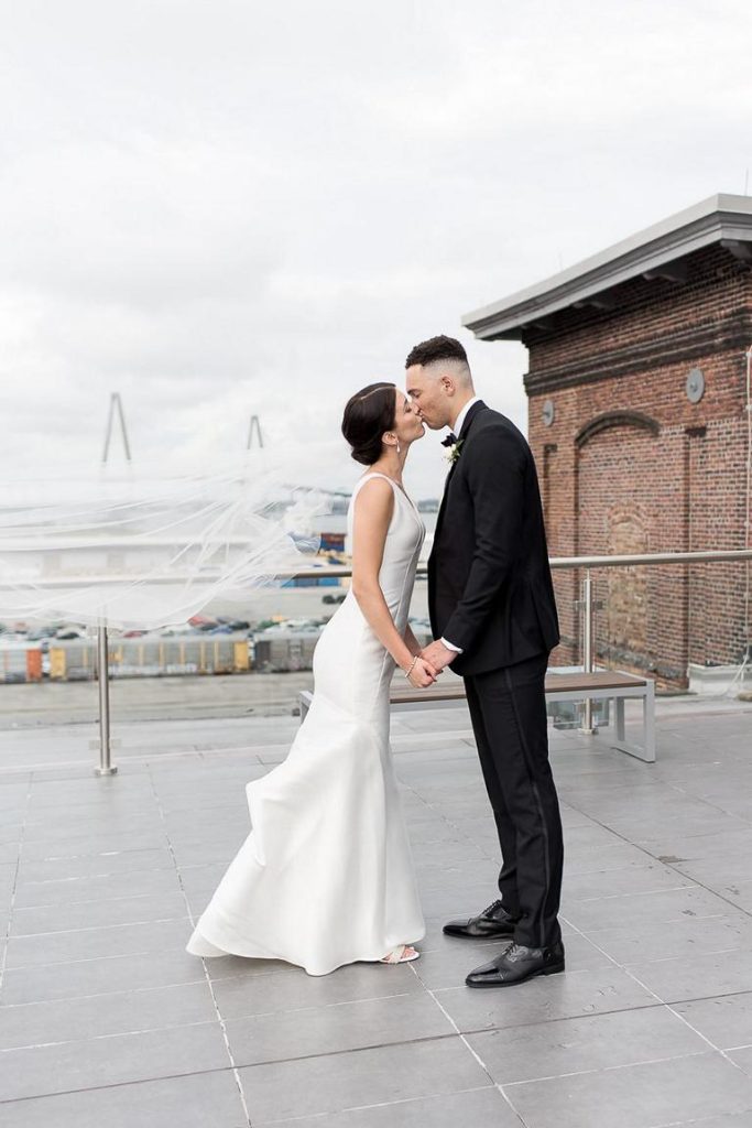 Bride and groom kiss on rooftop with bride's veil blowing in the wind