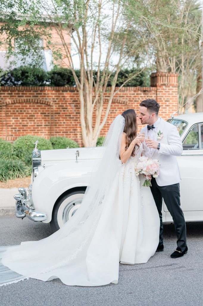 Newlyweds kiss and toast with champagne in front of vintage white Rolls Royce