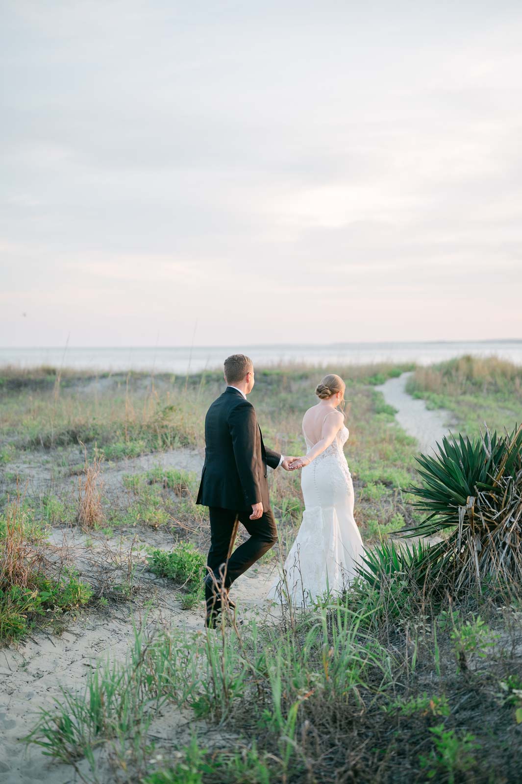 bride leads groom down beach path at sunset