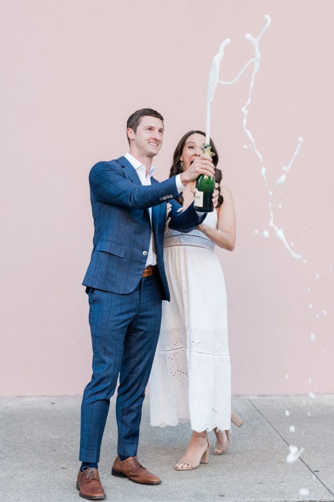 Newly engaged couple pop bottle of champagne in front of pink wall