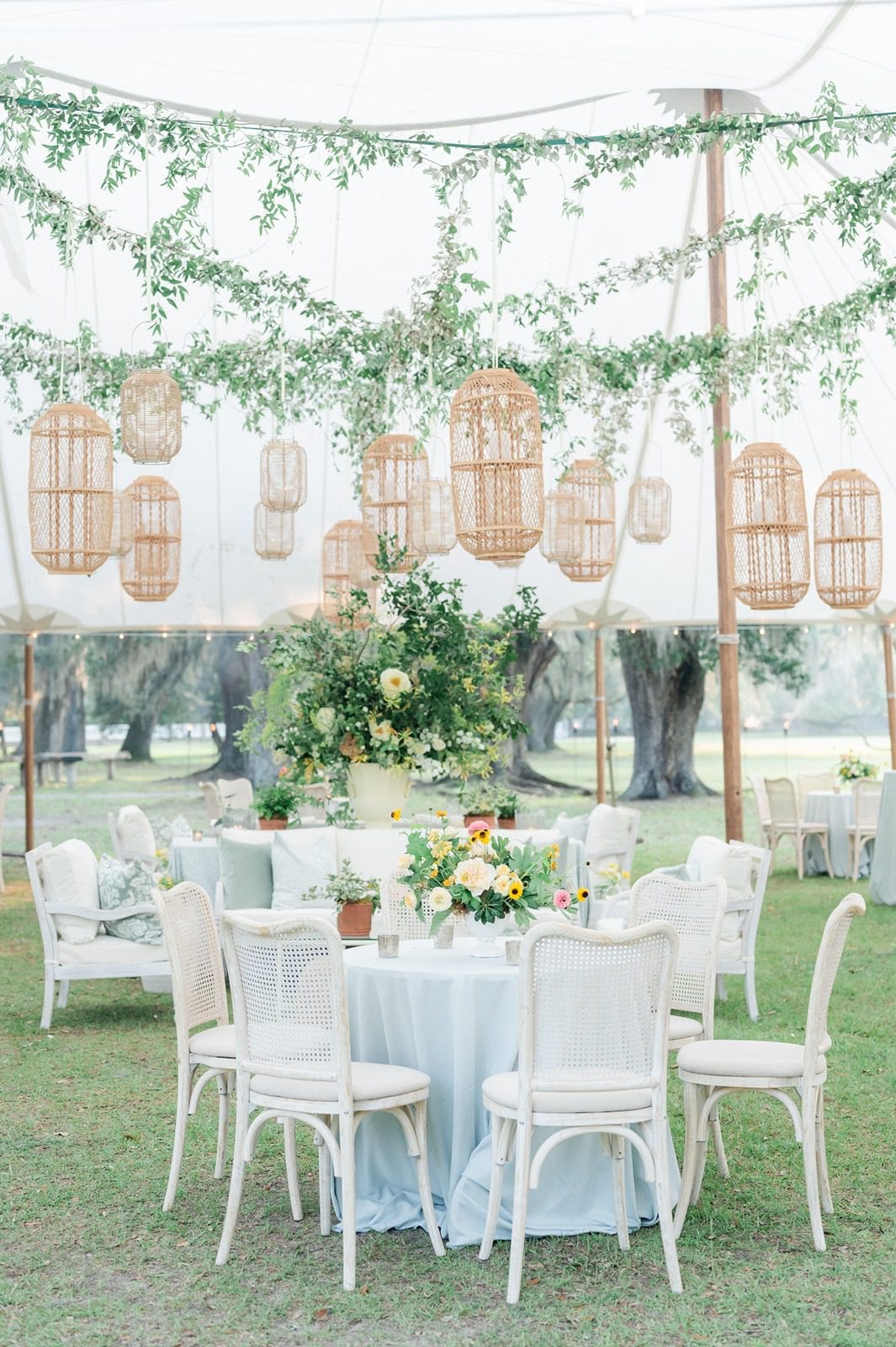 Wedding reception under tent with greenery strung from ceiling, white chairs and couches, rattan floating chandeliers