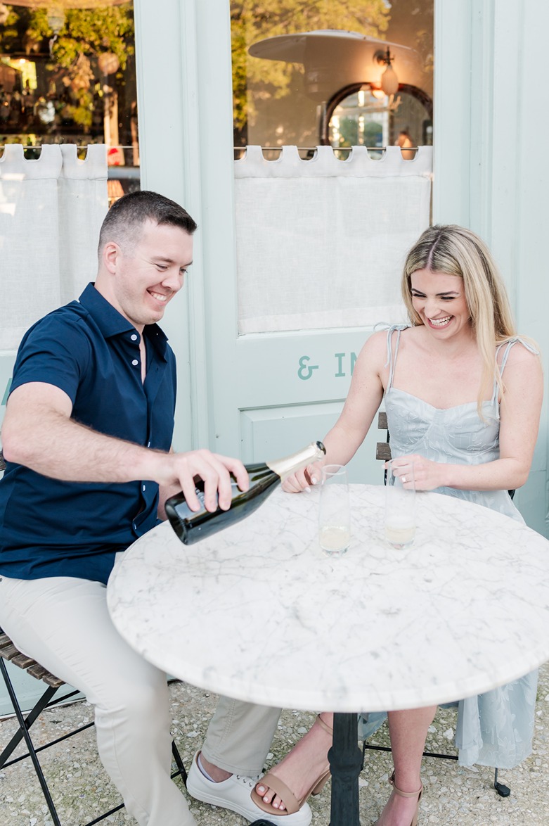 guy pours champagne at table while fiancée looks on
