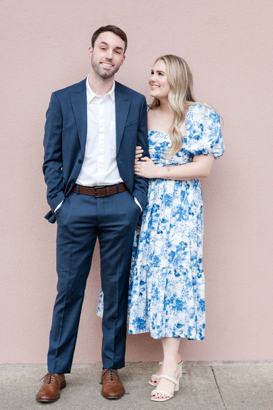 engaged couple stand in front of pink building with girl hanging on to guy's arm