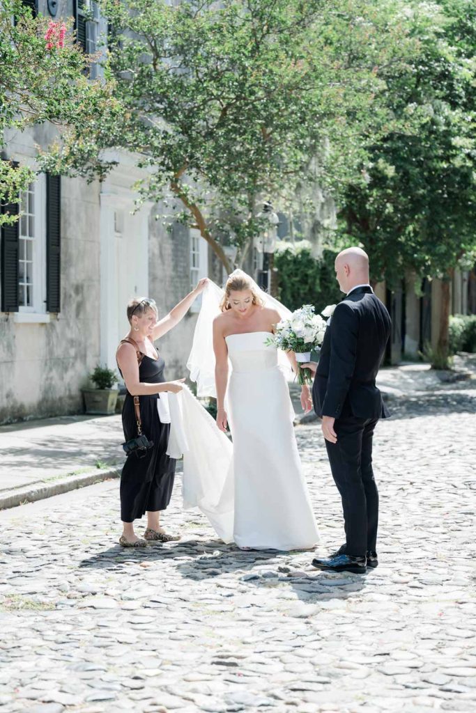 photographer helping bride with veil while groom looks on holding bouquet