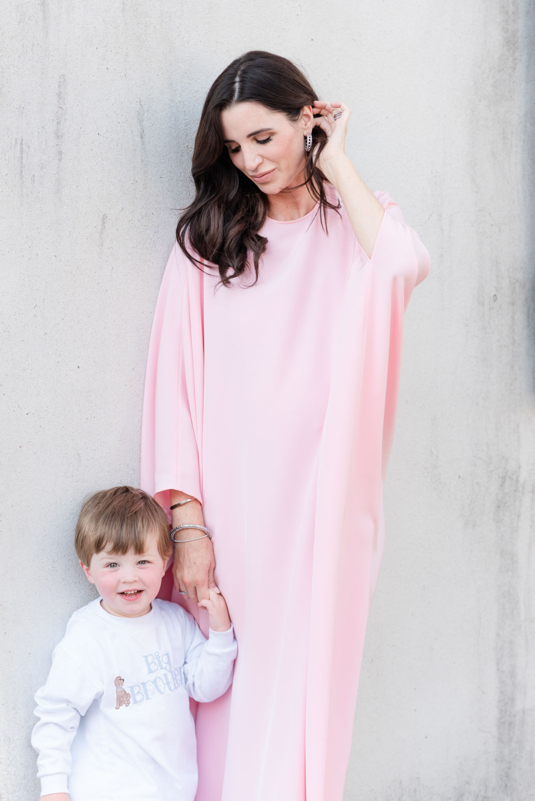 pregnant woman in pink kaftan tucking hair behind ear while holding toddler son's hand